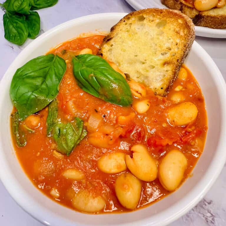 A white bowl filled with tomato and Marry Me butter bean soup garnished with fresh basil leaves. A slice of toasted bread is nestled in the bowl, while another plate with toast teases in the background.