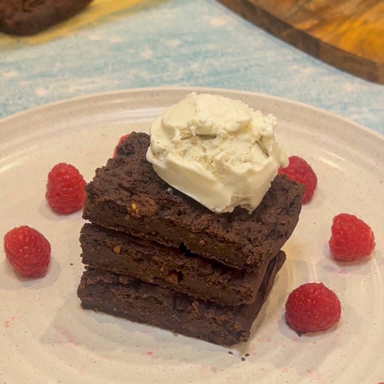 A stack of three sweet potato brownies topped with a scoop of vanilla ice cream sits on a white plate. Fresh raspberries are scattered around the brownies, and a light blue surface is partially visible beneath the plate.