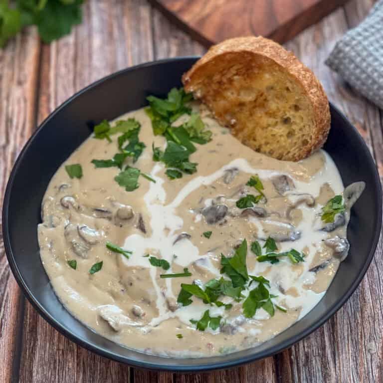 A rustic wooden table cradles a black bowl brimming with creamy mushroom soup. Garnished with fresh parsley and a swirl of cream, it invites you to savor each silky spoonful. A slice of toasted bread rests on the rim, completing this cozy culinary moment.