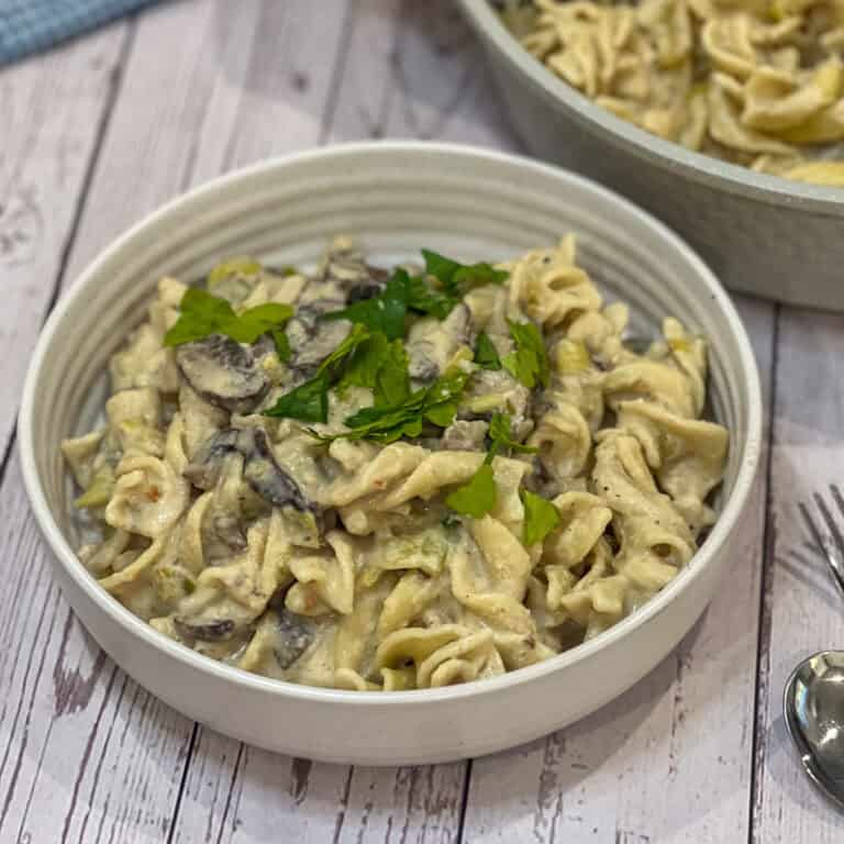 A white bowl filled with creamy leek and mushroom pasta is garnished with fresh parsley. The savory dish is set on a rustic wooden surface, accompanied by a fork and spoon.