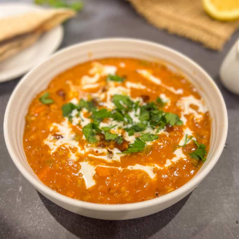 A bowl of rich, creamy dal makhani, reminiscent of a hearty red lentil soup, garnished with fresh cilantro and a drizzle of cream. The dish boasts a vibrant orange-red hue and smooth texture. In the background, there are flatbreads accompanied by a lemon slice on a plate.