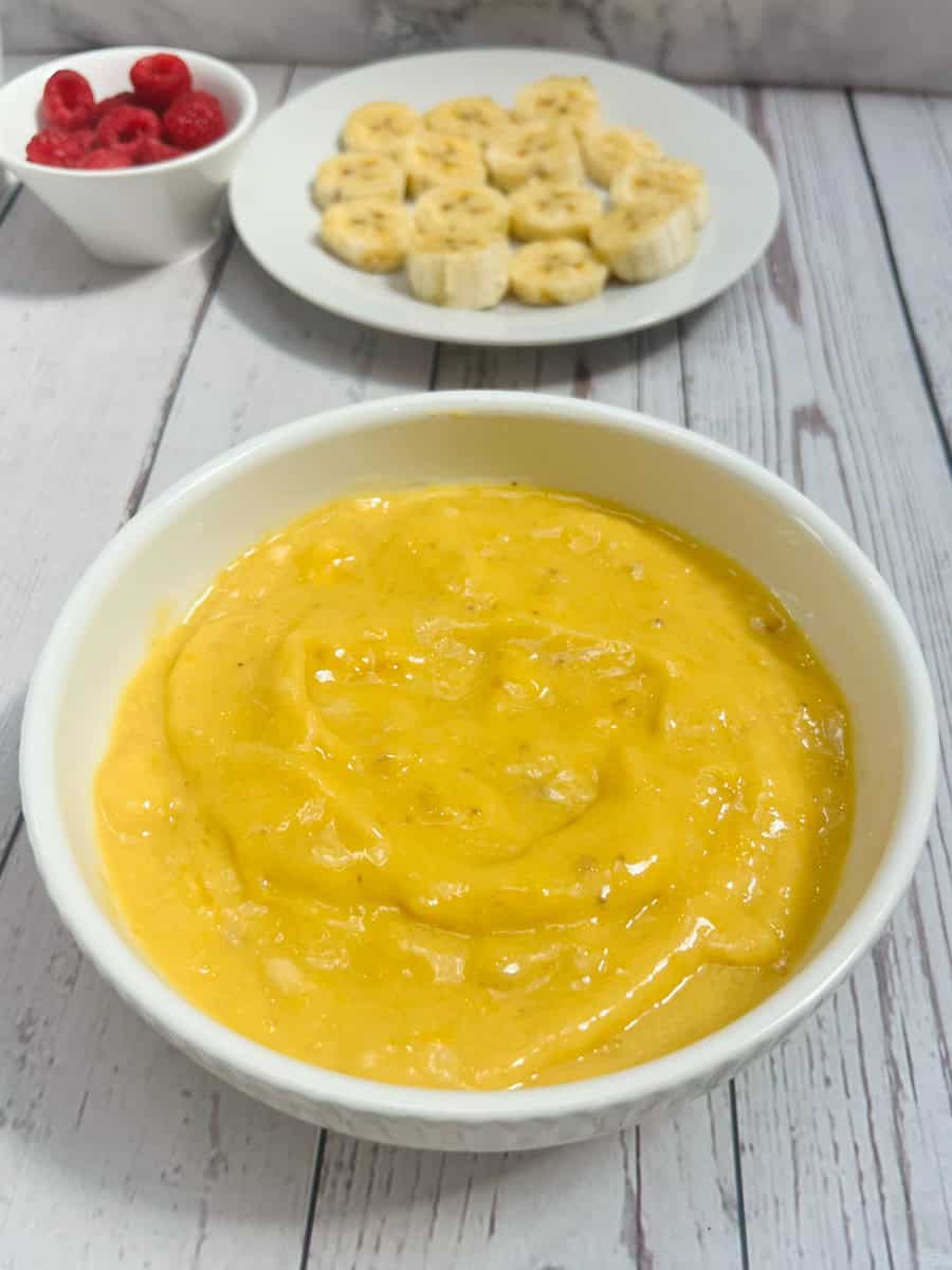 A bowl of smooth, creamy yellow pudding sits on a wooden surface. In the background, theres a plate of sliced bananas and raspberries, reminiscent of a vibrant mango smoothie bowl.