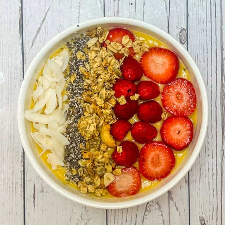 A vibrant Mango Smoothie Bowl topped with sliced strawberries, raspberries, granola, chia seeds, coconut flakes, and cashews sits invitingly on a white wooden surface.