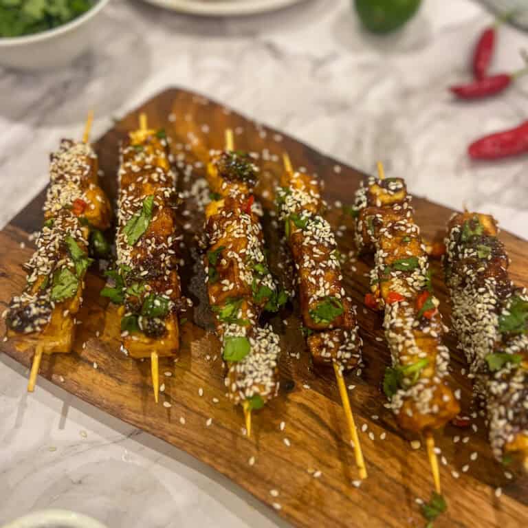 Wooden board with skewered grilled tofu pieces, drizzled with sauce, garnished with sesame seeds, herbs, and chopped red chili. Background shows scattered lime, herbs, and a bowl of greens.