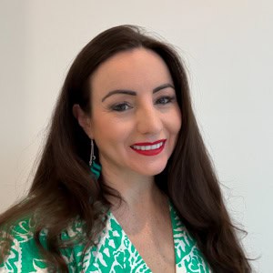 Woman with long brown hair and red lipstick smiles at the camera. She is wearing a green and white patterned top and turquoise earrings, standing against a plain background.