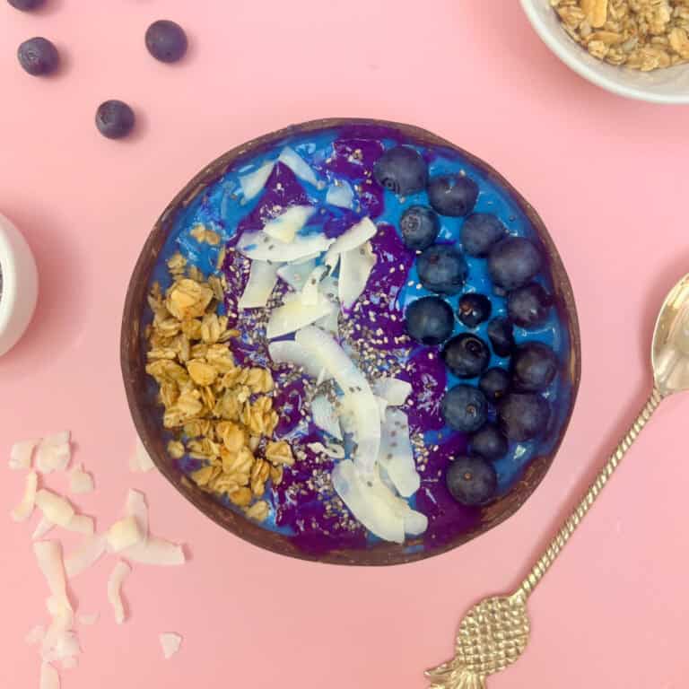 A vibrant blue spirulina smoothie bowl on a pink background, adorned with granola, coconut flakes, and juicy blueberries. Beside it rests a spoon with a decorative handle, while scattered blueberries and coconut flakes complete the enticing scene.