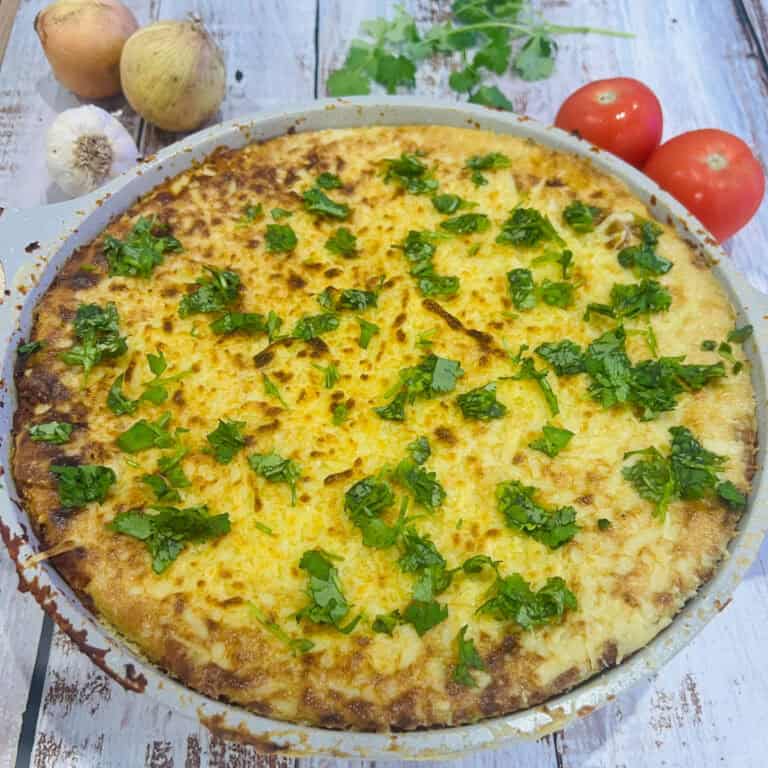 A vegan one-pot lasagne with a golden-brown crust topped with fresh cilantro. Surrounding this delightful dish are whole tomatoes, an onion, garlic, and cilantro on a rustic white wooden surface.