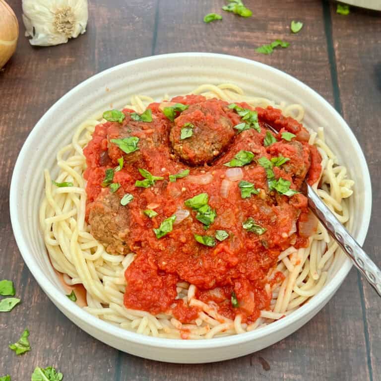 A bowl of spaghetti topped with red tomato sauce and vegan meatballs, garnished with chopped green herbs. The dish is on a wooden table with a fork placed to the side of the bowl.