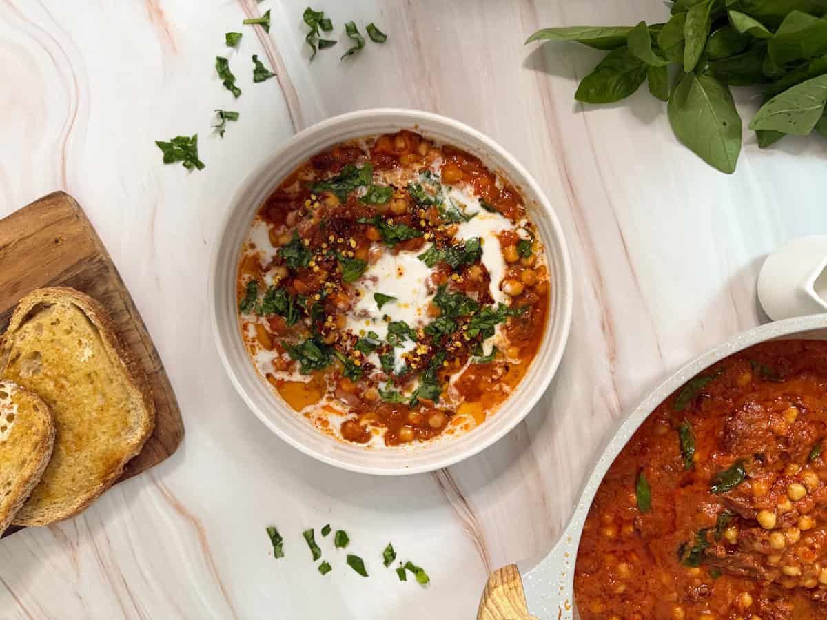 A bowl of chickpea stew with tomato sauce, garnished with cilantro and a drizzle of cream, sits on a marble surface. Slices of toasted bread rest on a wooden board nearby, and fresh spinach leaves are scattered on the counter.