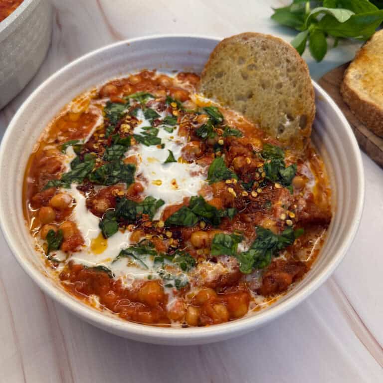 A white bowl filled with creamy tomato and chickpea soup, topped with yogurt, chopped herbs, and chili flakes. A slice of crusty bread is dipped into the soup, while fresh herbs are seen in the background.