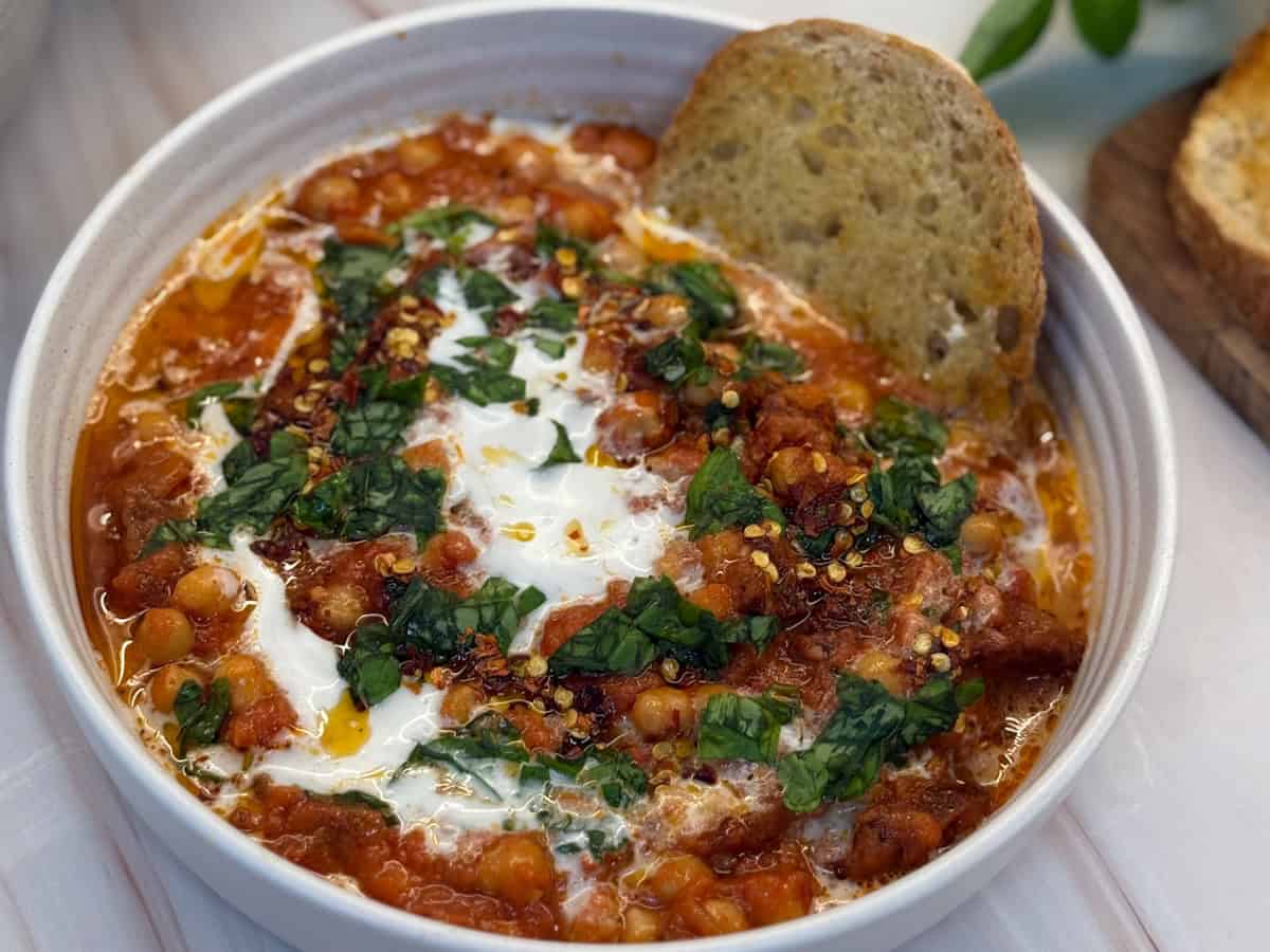 A bowl of spicy chickpea stew garnished with a swirl of yogurt, chopped herbs, and red pepper flakes. A slice of crusty bread is placed on the edge of the bowl.