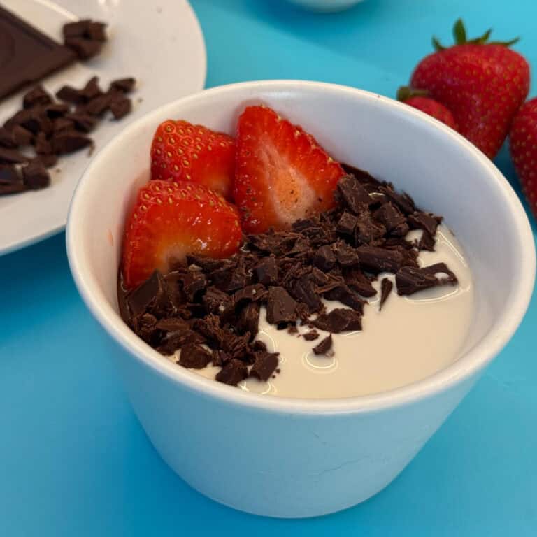 A white bowl filled with creamy yogurt topped with sliced strawberries and dark chocolate shavings evokes the indulgence of avocado chocolate mousse. In the background, whole strawberries and a plate with dark chocolate pieces sit enticingly on a blue surface.