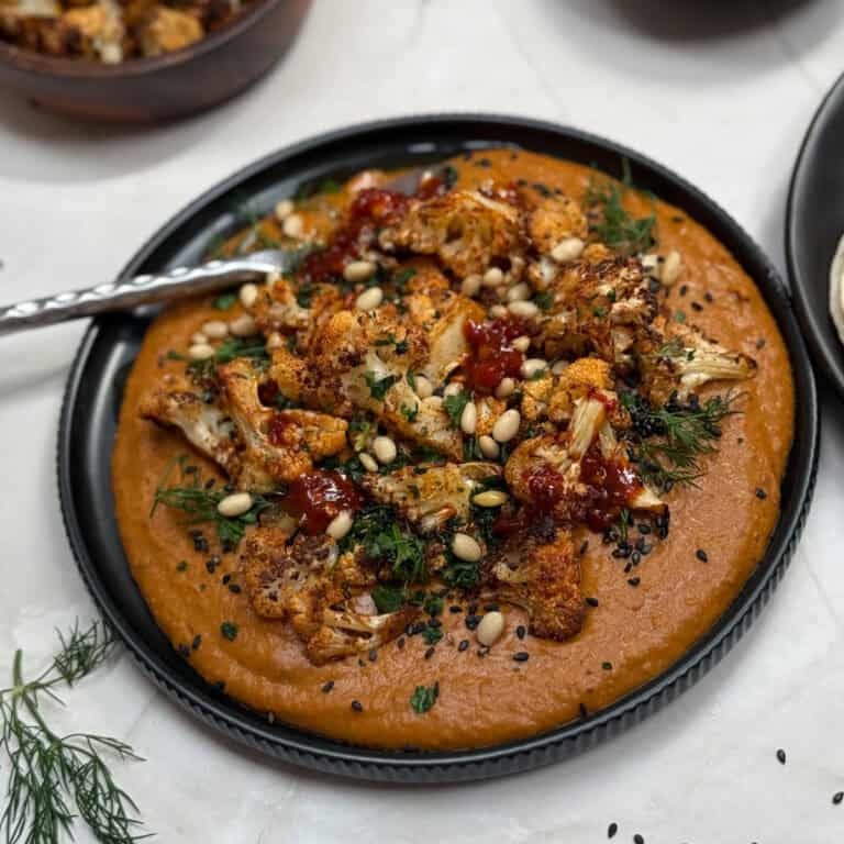 A black plate with creamy hummus topped with roasted cauliflower, pine nuts, black sesame seeds, fresh herbs, and a drizzle of spicy harissa. A silver spoon rests on the side of the dish.