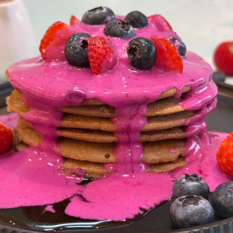 A stack of buckwheat pancakes topped with vibrant pink sauce, blueberries, and halved strawberries on a dark plate. More berries are scattered around, creating a colorful and appetizing display.