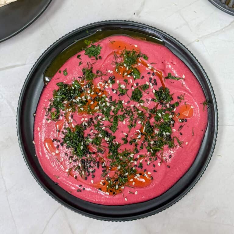 A black plate features vibrant pink beet hummus, subtly echoing a Beetroot and Coconut Yoghurt Dip. Its garnished with a drizzle of olive oil, black and white sesame seeds, and finely chopped fresh herbs, elegantly set on a crisp white surface.