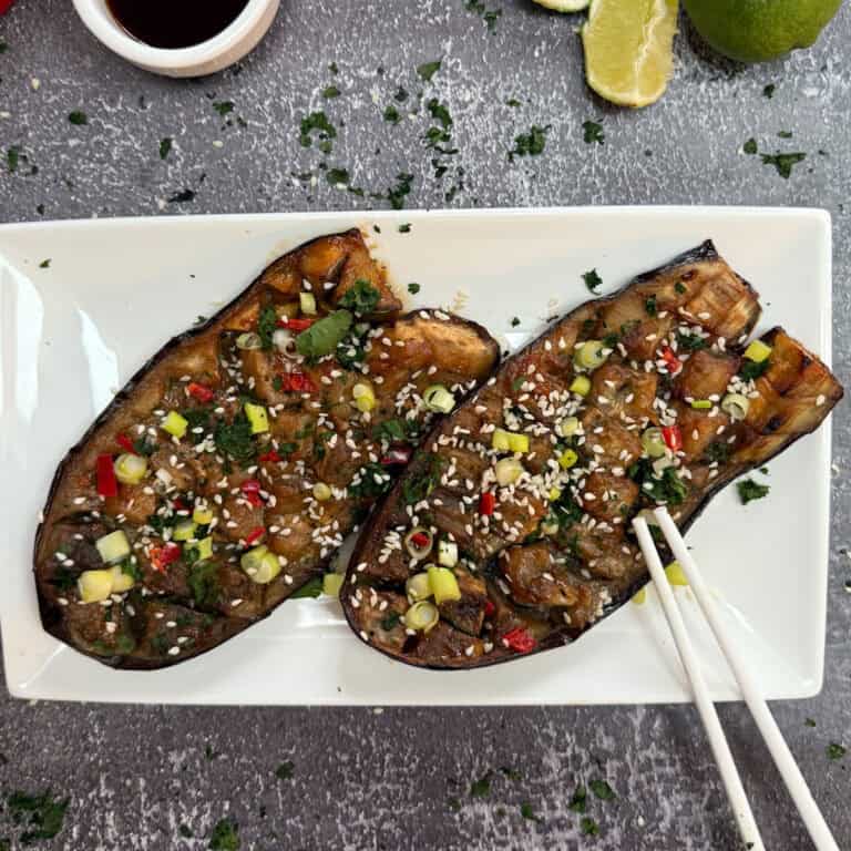 Two miso glazed aubergine halves rest on a rectangular white plate, topped with diced vegetables, sesame seeds, and herbs. Chopsticks accompany the dish. A lime and a small bowl of sauce sit nearby, all set against a dark textured surface.