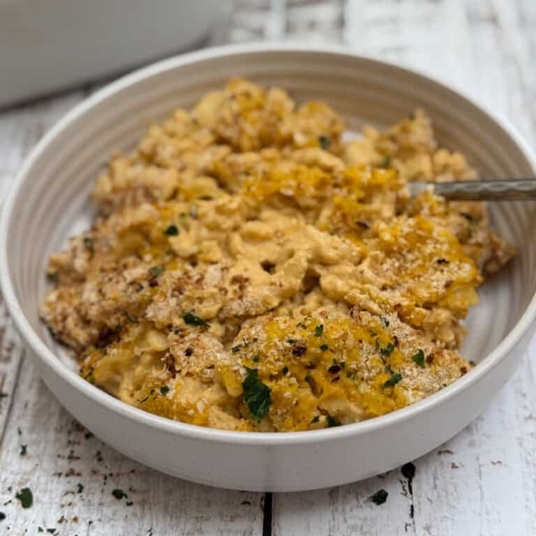 A round white dish contains baked vegan mac n cheese, topped with golden breadcrumbs and garnished with parsley. The creamy pasta is on a rustic white wooden table, with a metal serving spoon visible on the side.