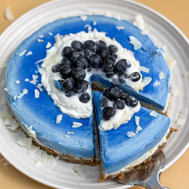 A sliced blue cashew cheesecake on a white plate, topped with whipped cream and fresh blueberries. Coconut flakes are scattered on top and around the plate. One slice is being removed with a serving spatula.