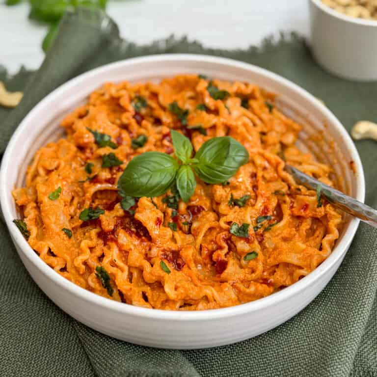 A white bowl filled with creamy, ruffled pasta coated in rich Cashew and Red Pepper sauce, garnished with fresh basil leaves. The bowl sits on a dark green cloth with a fork resting on the edge.