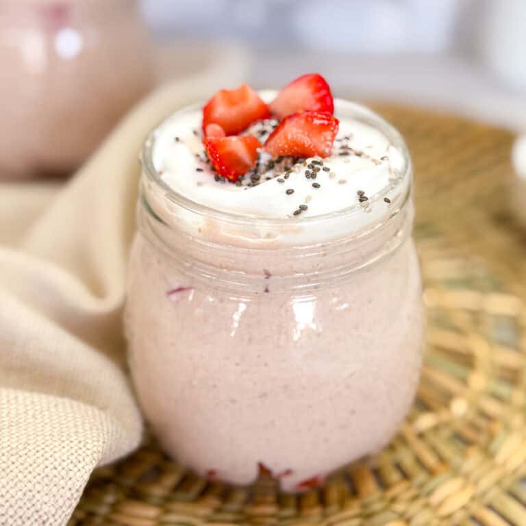A jar filled with creamy chia pudding and strawberry chunks, topped with chia seeds, sits on a woven mat. Beside it, a beige cloth is draped elegantly. Enjoy this delightful treat paired with a bowl of strawberry blended oats for the perfect breakfast duo.