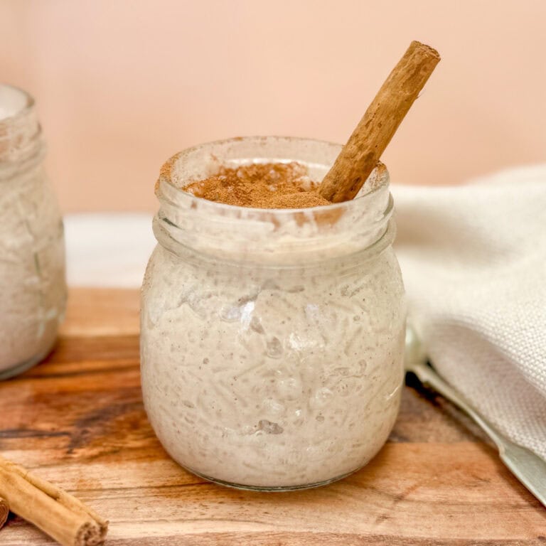 A glass jar filled with creamy vegan rice pudding topped with ground cinnamon and a cinnamon stick. Another jar is partially visible in the background on a wooden surface with a white fabric nearby.