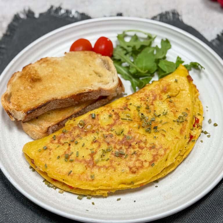 A plate featuring a golden omelette sprinkled with herbs, accompanied by two slices of toasted bread. Fresh arugula and two cherry tomatoes are on the side, all set on a textured black placemat.