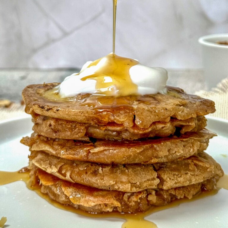 A stack of pancakes topped with a dollop of cream and syrup being drizzled over. The pancakes are on a white plate, and a cup is visible in the blurred background.