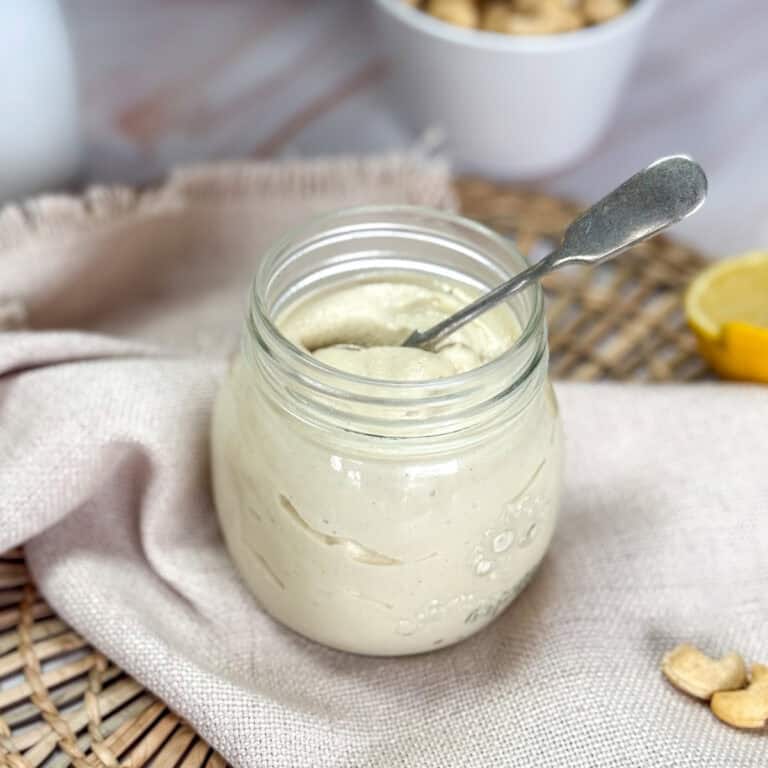 A glass jar filled with creamy, smooth cashew butter is placed on a beige cloth. A spoon is dipped into the jar. Background elements include a white bowl with cashews and a lemon wedge. The arrangement rests on a woven mat with blurred wooden texture.