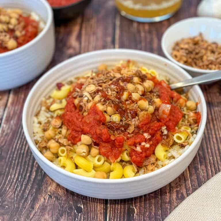 Bowl of koshari with rice, macaroni, chickpeas, and fried onions, topped with tomato sauce. The dish is set on a wooden table, with additional bowls of ingredients around it.