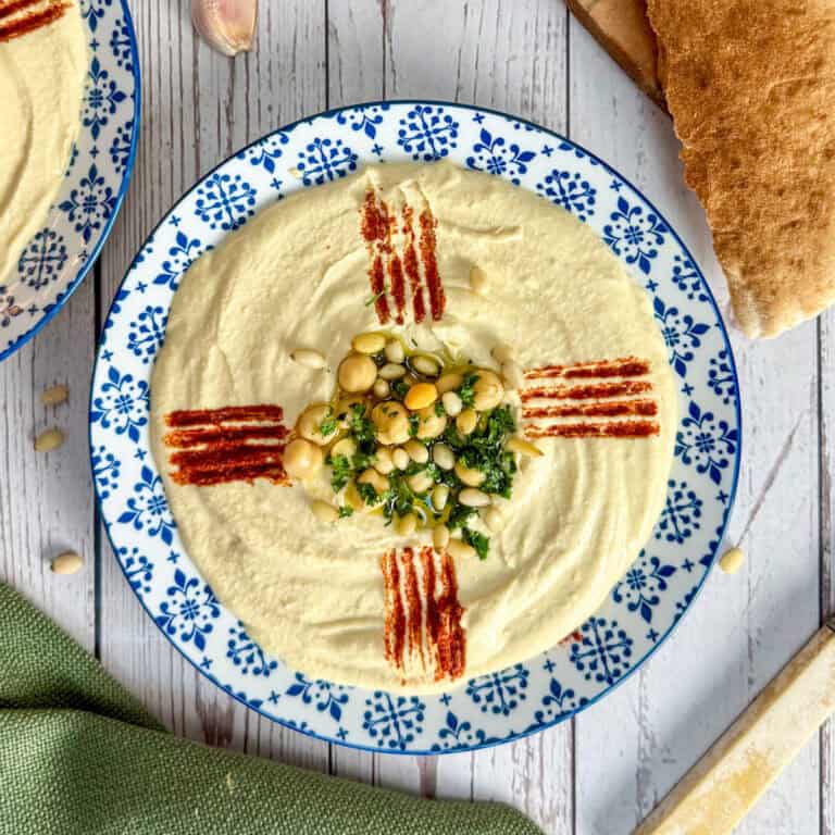 A plate of creamy hummus garnished with whole chickpeas, chopped parsley, pine nuts, and lines of paprika, served with pita bread on a white wooden table.