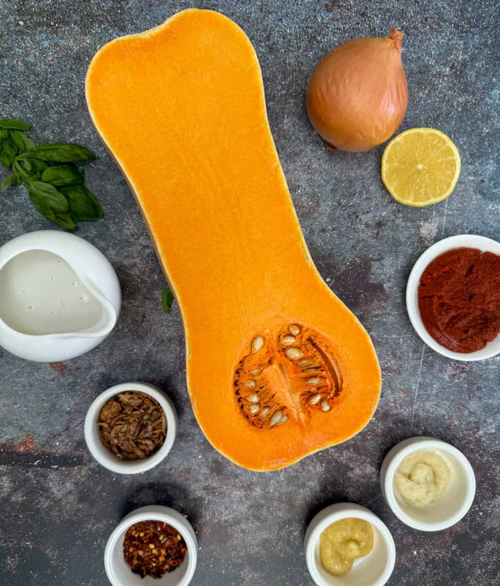 A halved butternut squash surrounded by an onion, half a lemon, fresh basil, a small pitcher of cream, tomato paste, and small bowls of spices and seasonings on a dark countertop.