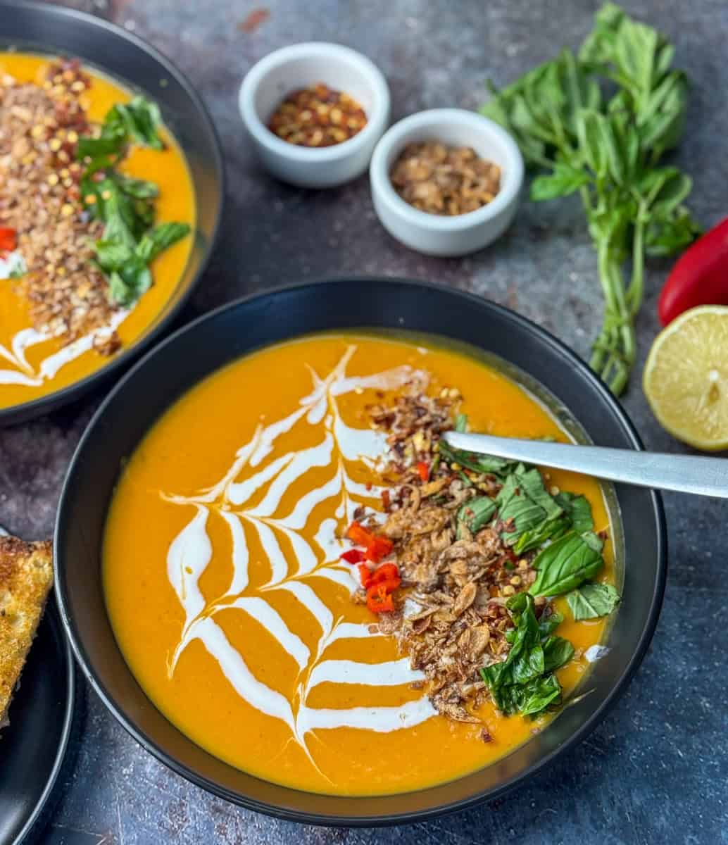 A bowl of orange soup garnished with coconut milk in a spiderweb pattern, herbs, chili, crispy onions, and seeds, with small bowls of spices, fresh basil, a red chili, and a lemon beside it.