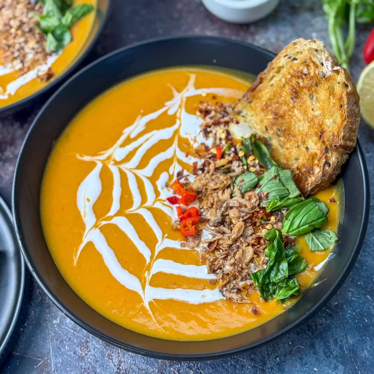 A bowl of creamy orange soup garnished with a white swirl, crispy bits, chopped red chili, fresh basil, and served with a slice of toasted bread. The soup is presented in a black bowl on a dark surface.