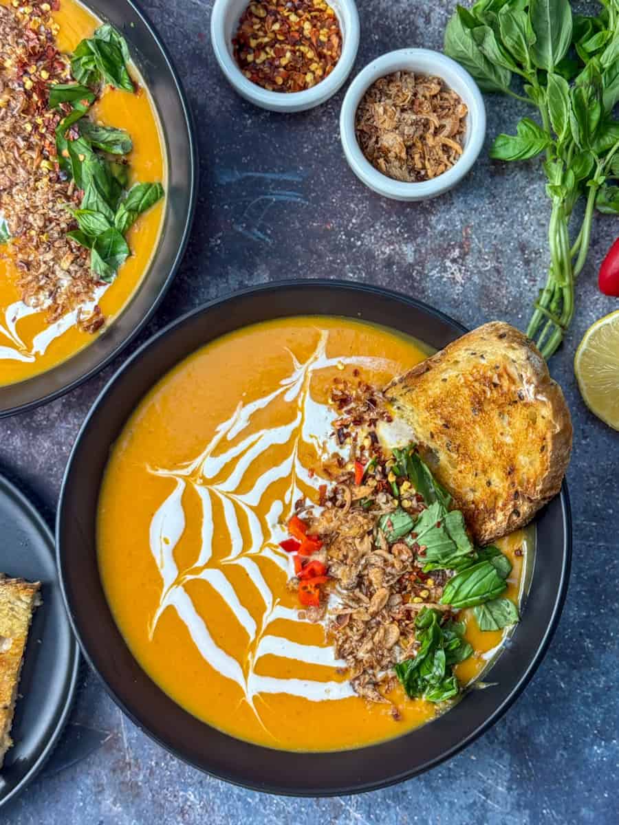 A bowl of creamy orange soup with a white spiderweb design, garnished with herbs and fried onions, served with toasted bread. Small bowls of chili flakes and toppings, basil, and a lemon are nearby.