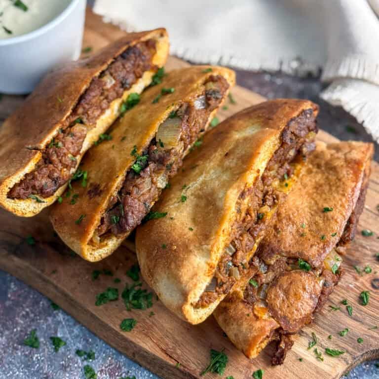 Three stuffed pita pockets filled with a savory mixture of ground meat, onions, and herbs are arranged on a wooden board, garnished with chopped parsley, with a bowl of white sauce in the background.