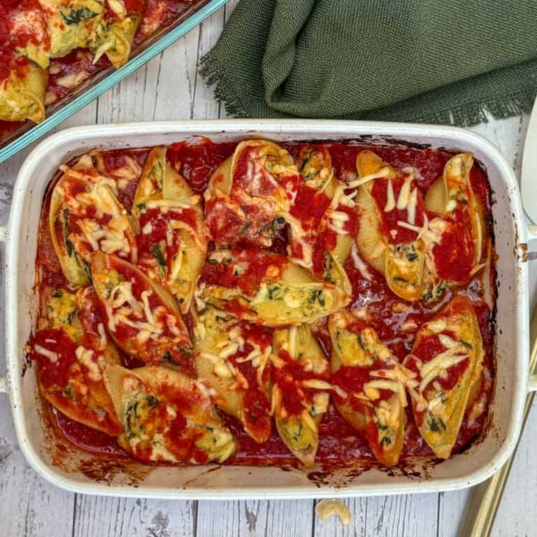 A baking dish filled with large pasta shells stuffed with a spinach and cheese mixture, topped with tomato sauce and melted cheese, sitting on a white wooden surface with a green cloth nearby.
