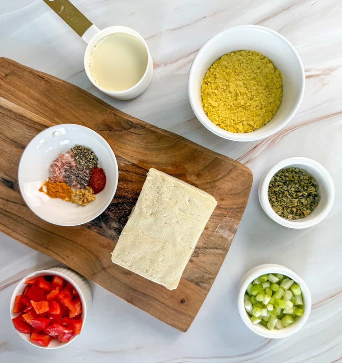A block of tofu on a wooden board surrounded by small bowls of chopped red bell pepper, green onions, dried herbs, spices, nutritional yeast, and a measuring cup of plant-based milk on a marble surface.