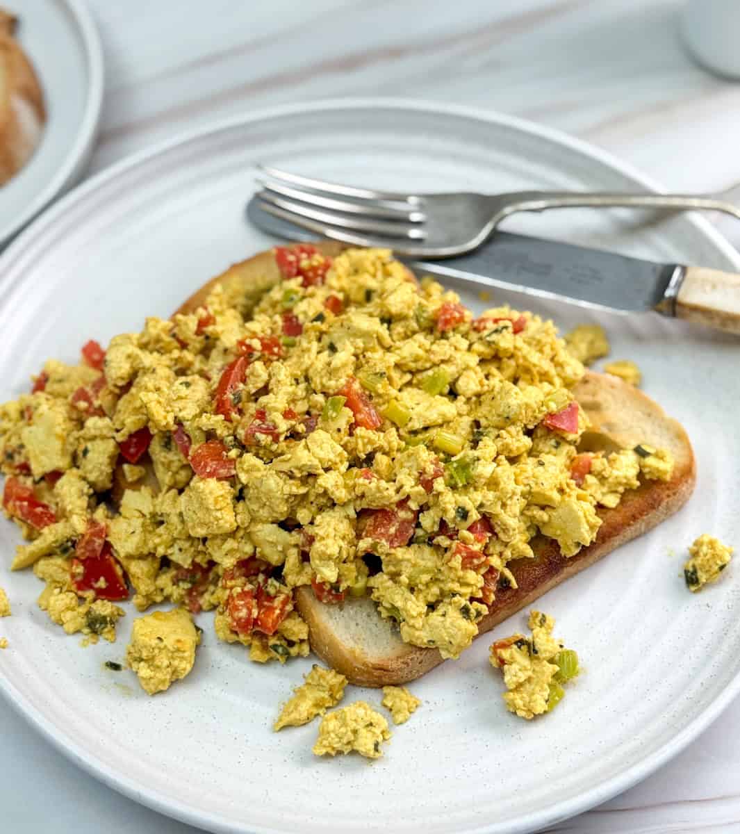 A plate with scrambled tofu mixed with chopped tomatoes, green vegetables, and spices, served on a slice of toasted bread. A fork and knife rest on the plate in the background.