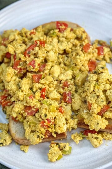 Scrambled tofu mixed with red and green peppers served on two slices of toasted bread on a white plate, with a fork placed beside the toast.