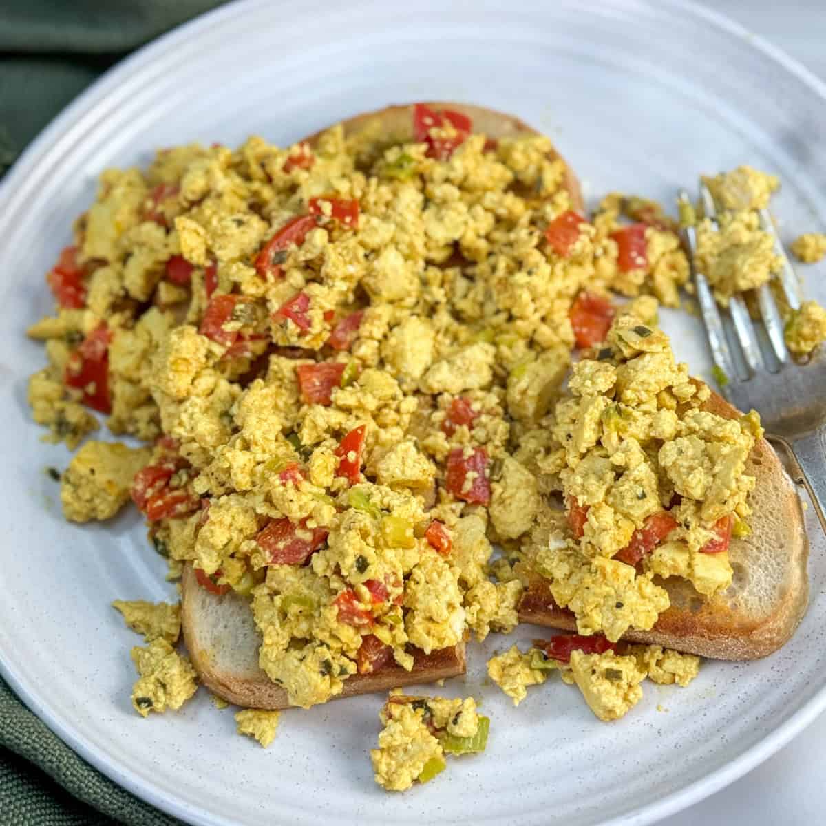 A plate with two slices of toast topped with scrambled tofu mixed with diced red peppers and green onions, with a fork resting on the plate.