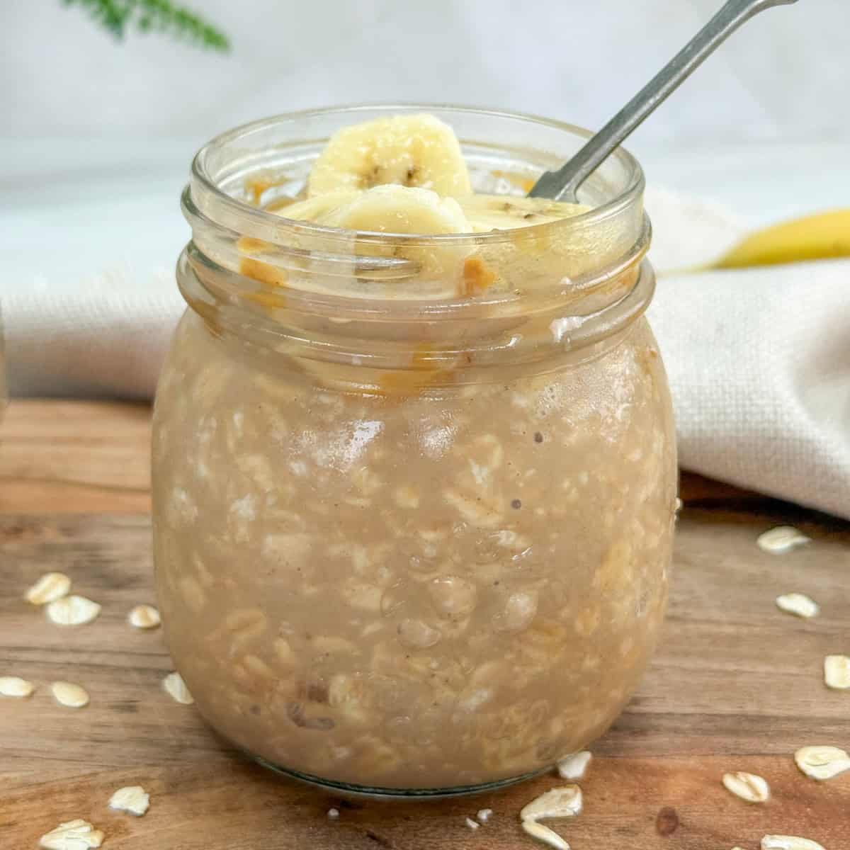 A glass jar filled with overnight oats topped with banana slices and a drizzle of caramel, with a spoon inside. Some oats are scattered on the wooden surface next to the jar.