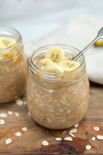 Two glass jars filled with overnight oats topped with banana slices, placed on a wooden surface with scattered oats and a spoon in one jar. A beige cloth and banana are in the background.