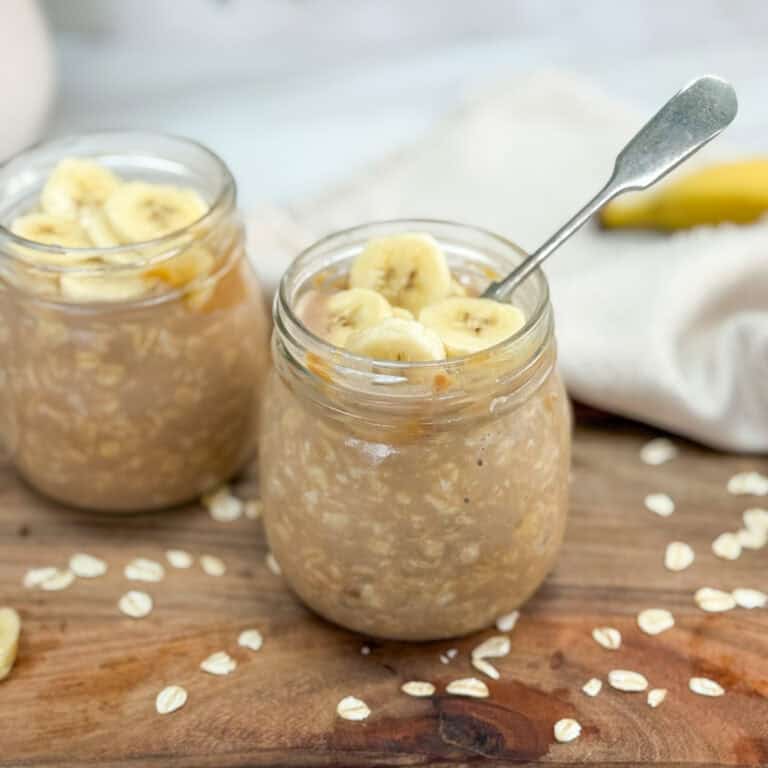 Two glass jars filled with overnight oats topped with banana slices, placed on a wooden surface with scattered oats and a spoon in one jar. A beige cloth and banana are in the background.