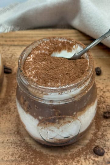 A glass jar filled with creamy tiramisu, topped with cocoa powder. A spoon rests inside the jar, and some cocoa powder is sprinkled on the wooden surface around the jar. Coffee beans are also visible nearby.
