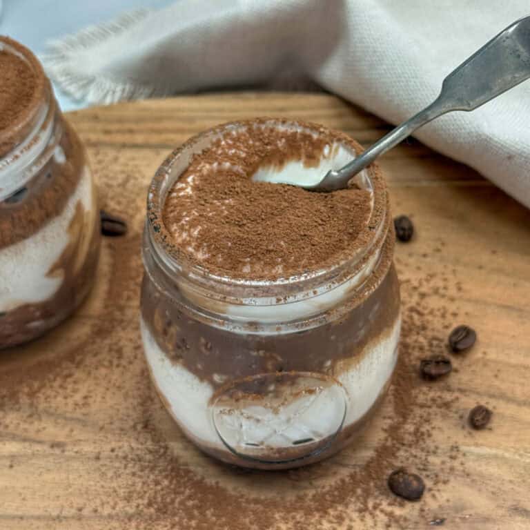 A glass jar filled with creamy tiramisu, topped with cocoa powder. A spoon rests inside the jar, and some cocoa powder is sprinkled on the wooden surface around the jar. Coffee beans are also visible nearby.