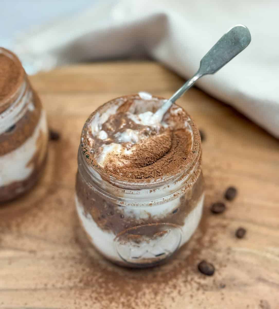 A jar filled with creamy dessert layered with cocoa powder, possibly tiramisu or a mousse, sits on a wooden surface. A spoon rests inside the jar, and some cocoa powder and coffee beans are scattered nearby.