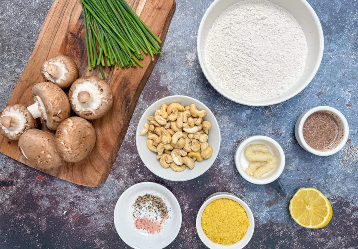A top-down view of cooking ingredients: mushrooms and chives on a wooden board, and bowls with flour, cashews, ground flaxseed, minced garlic, lemon half, nutritional yeast, and seasonings on a dark countertop.