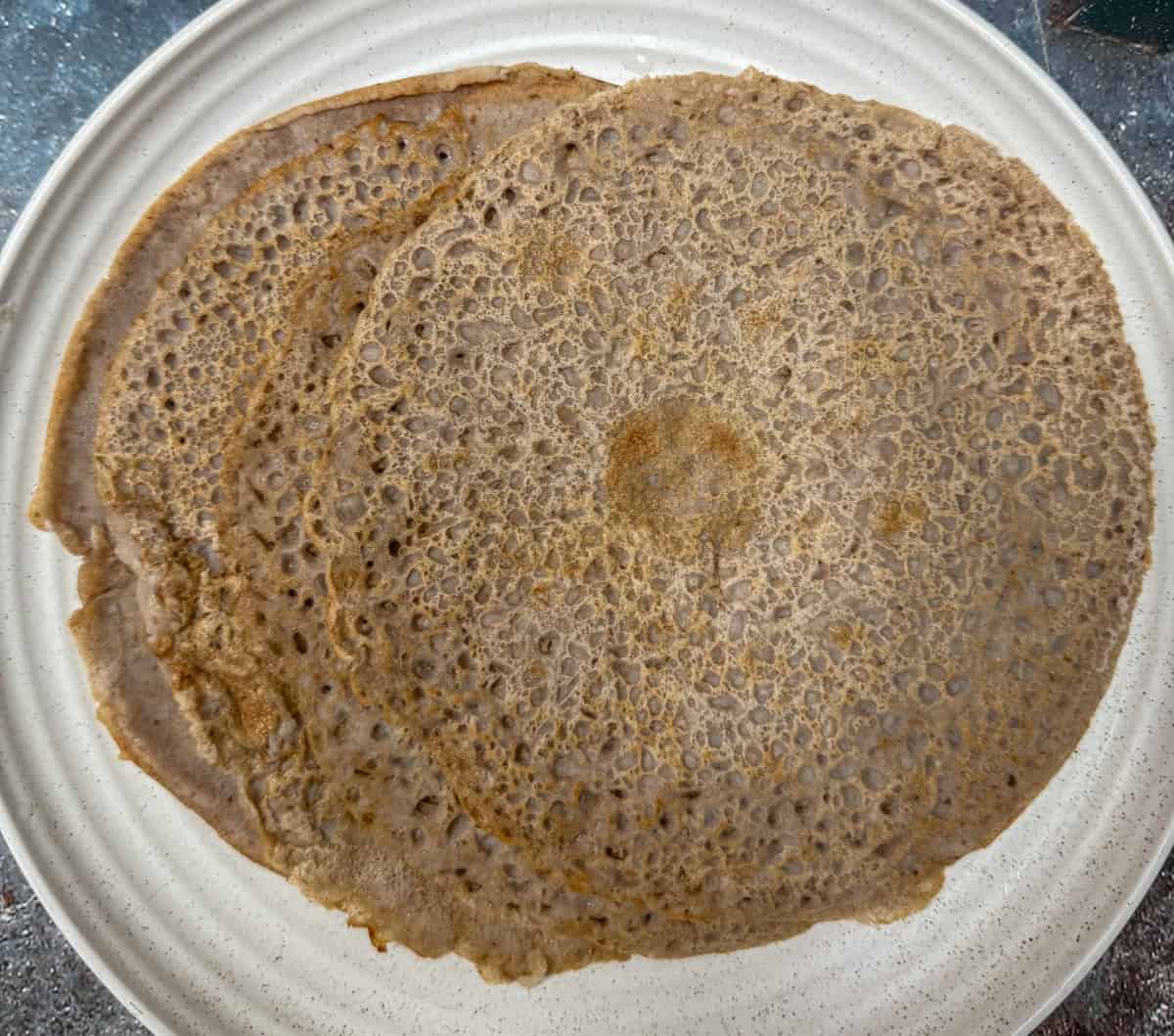 Two round, thin, brown pancakes with a lacy texture are placed on a white, ridged plate. The pancakes overlap slightly and have uneven, rustic edges.