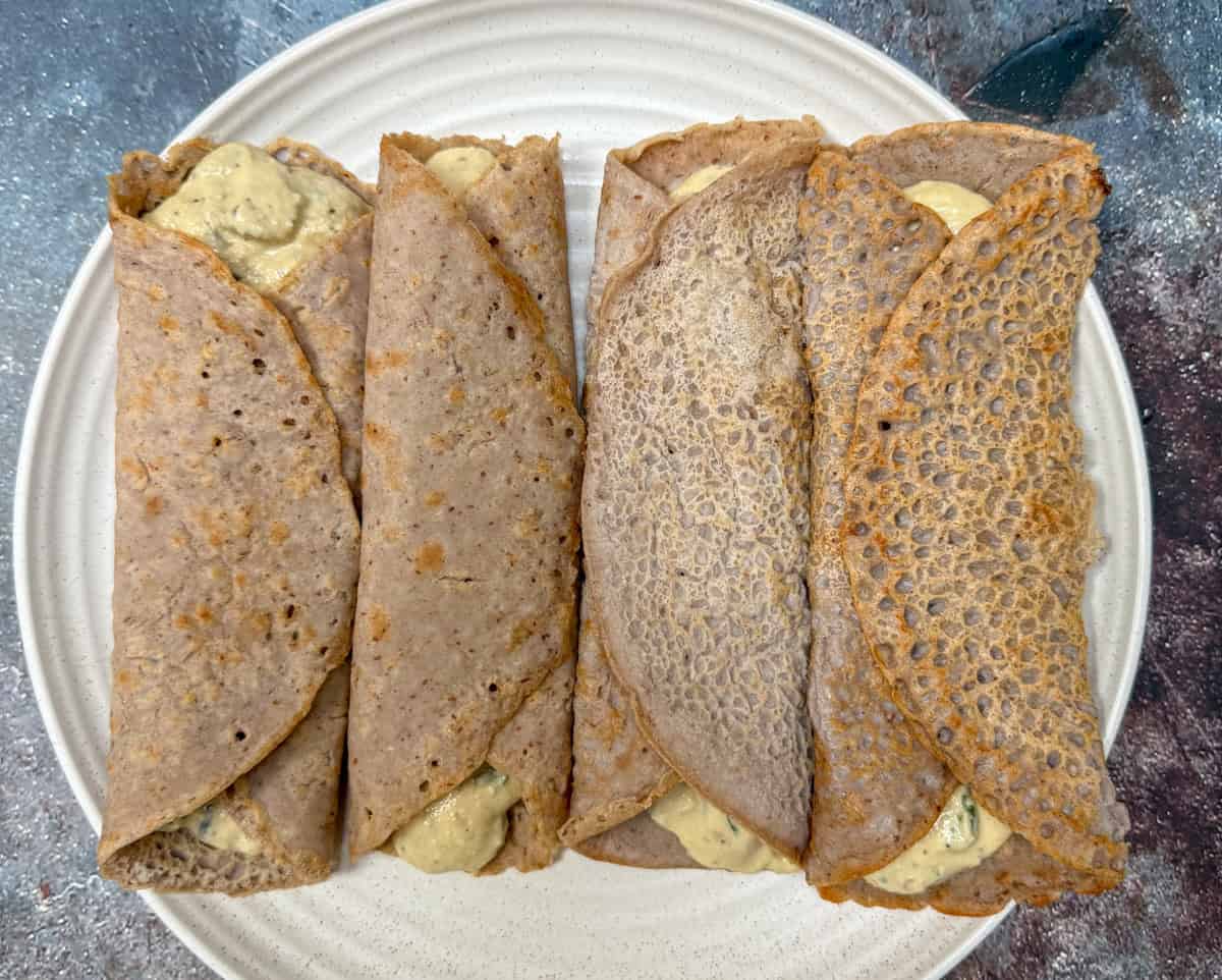 Four rolled buckwheat crepes filled with a creamy, savory mixture are arranged side by side on a white plate, placed on a textured surface.