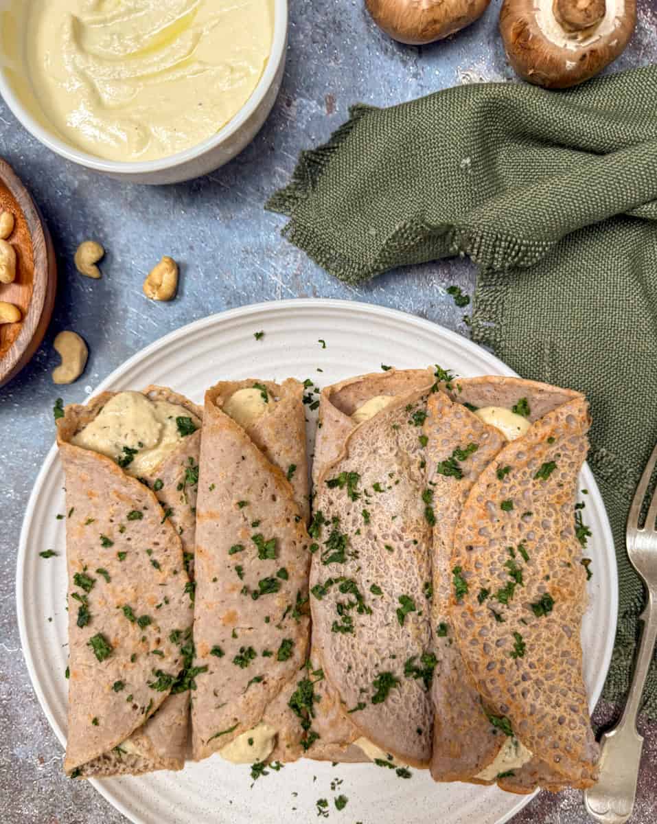 Four folded buckwheat crepes filled with a creamy filling and topped with chopped herbs are arranged on a white plate. A green cloth, cashews, mushrooms, and a bowl of creamy spread are nearby.